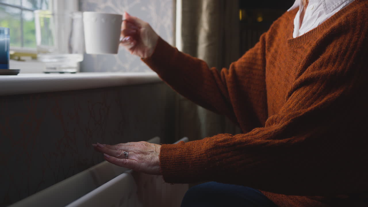 Primo piano di donna anziana con bevanda calda che cerca di riscaldarsi accanto al termosifone a casa durante la crisi energetica
