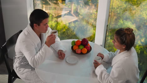 couple drinks coffee near window at table. woman and man wearing gowns have breakfast together in gampling house. newlyweds happy being together