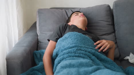 uncomfortable child sleeping covered in blanket adjusting on couch for comfort with tissue nearby, showing signs of discomfort during rest in peaceful indoor setting