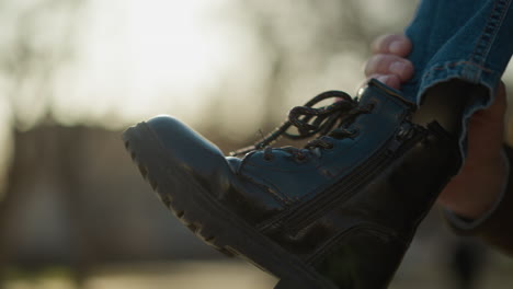 a close-up shot of a child's leg wearing blue jeans and a black boot. the child is being held up, with the background featuring an outdoor setting with trees and sunlight