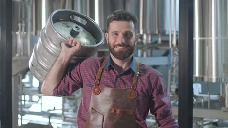 young brewer wearing a leather apron carries a barrel of craft beer at a modern brewery