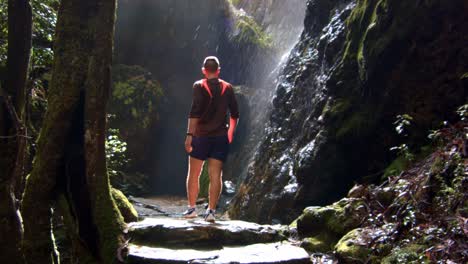 Adventurous-man-walking-into-rocky-cave,-water-flowing-down-the-rocks,-Springbrook-national-park,-Australia