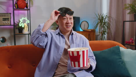 asian man sits on sofa eating popcorn and watching interesting tv serial, sport game online at home