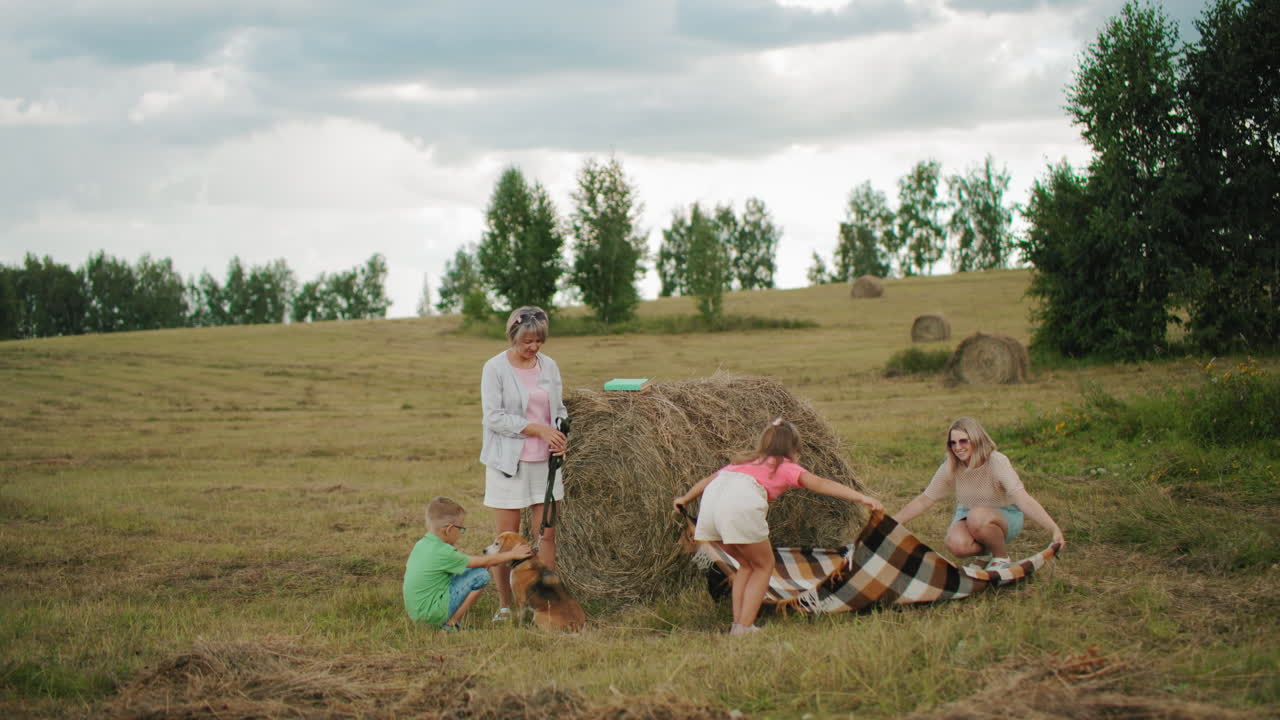 мать и дочь разложили коврик возле сенобоя на обширной сельскохозяйственной земле, бабушка стоит рядом, в то время как маленький мальчик касается собаки, создавая теплую, семейную сельскую сцену, наполненную связью.
