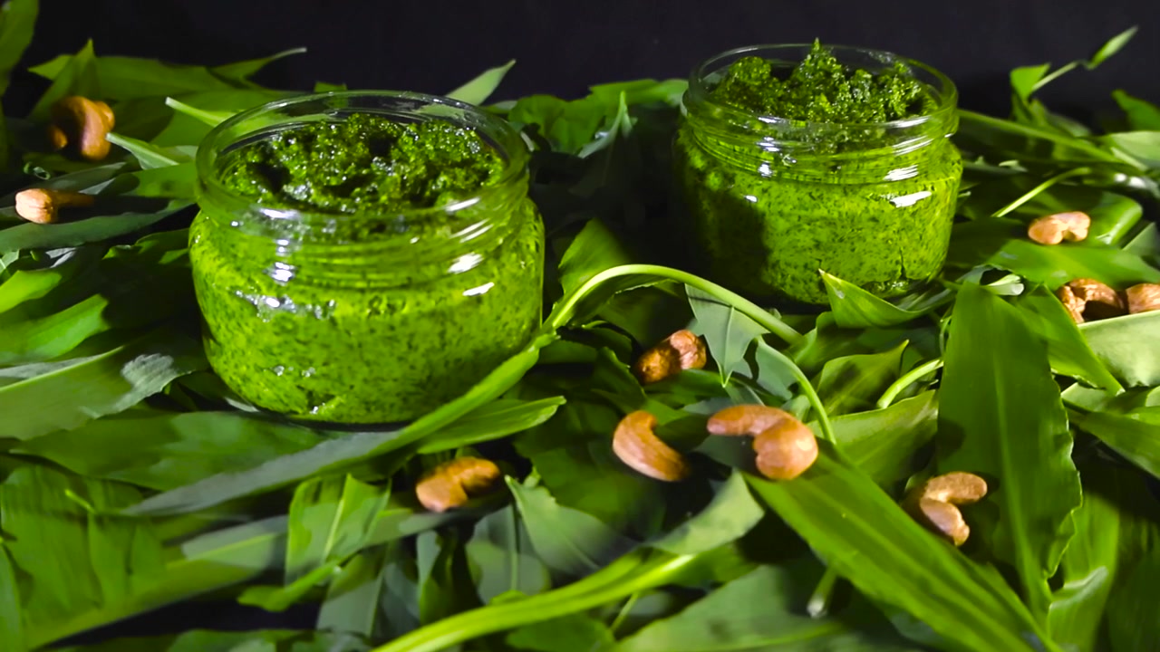 Siyah bir arka plan üzerinde dönen, taze roka, yabani sarımsak yaprakları ve karışık kuruyemişlerle çevrili, sağlıklı, vegan ve lezzetli bir sosu gösteren iki cam kavanoz ev yapımı yabani sarımsak pestosu.