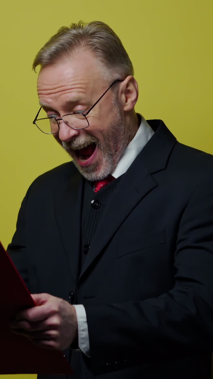 Happy elderly businessman with folder. Grey-bearded senior man is writing on paper on yellow background. Positive emotions. Vertical video
