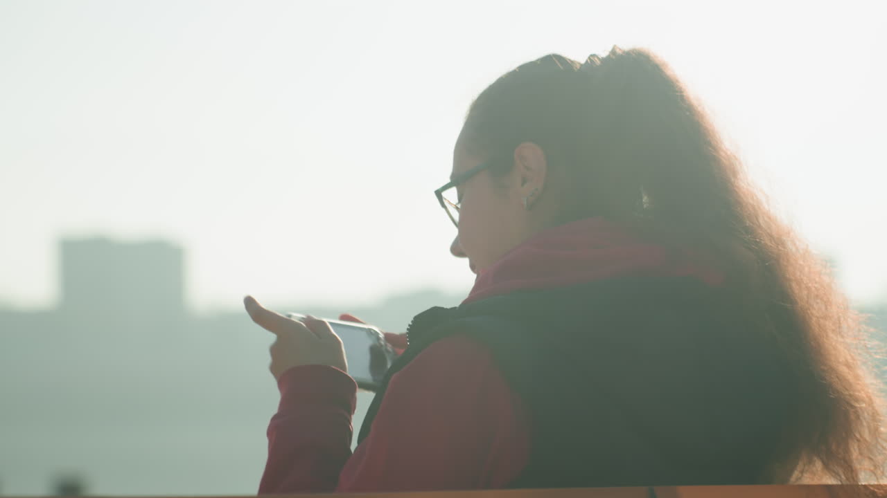 川沿いのベンチでスマートフォンを使うラティーナの女性。ゴールデンアワーの逆光でスカイラインを背景にしたポートレート。メッセージを確認したり、写真を構図したり、パノラマ撮影のために携帯を上げたりしている。眼鏡をかけ、ポニーテールで、赤いパーカーを着ている。