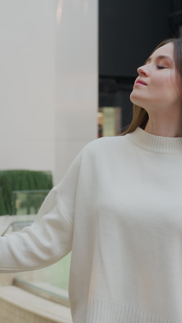 Woman in white sweater admires Christmas tree with glass-bordered water spring and decorative plant, urban structures in background, showcasing holiday ambiance in shopping mall