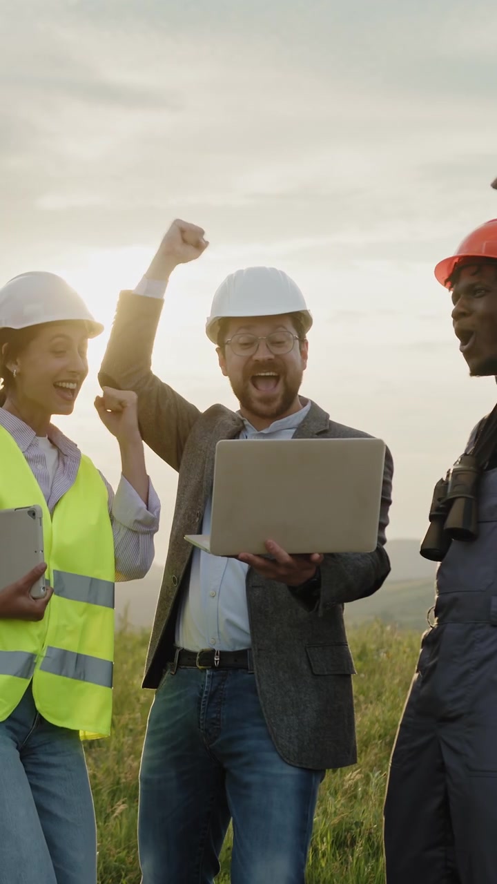 Diverse construction professionals celebrate achievement with laptops and tablets in a sunlit field