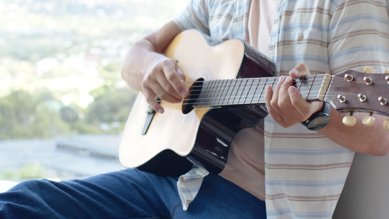 La mano derecha empieza a rasguear mientras el guitarrista se ajusta en la guitarra acústica, practicando cerca de una ventana.