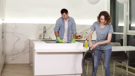 Cleaning-day.-Young-couple-cleaning-their-apartment---man-in-gloves-wiping-up-kitchen-stove,-girl-cleaning-floor-using-vacuum-cleaner.-Then-stop-and-smiling-to-the-camera