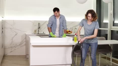 Día-De-Limpieza.-Pareja-Joven-Limpiando-Su-Apartamento---Hombre-Con-Guantes-Limpiando-La-Estufa-De-La-Cocina,-Niña-Limpiando-El-Piso-Usando-Una-Aspiradora.-Luego-Para-Y-Sonríe-A-La-Cámara.