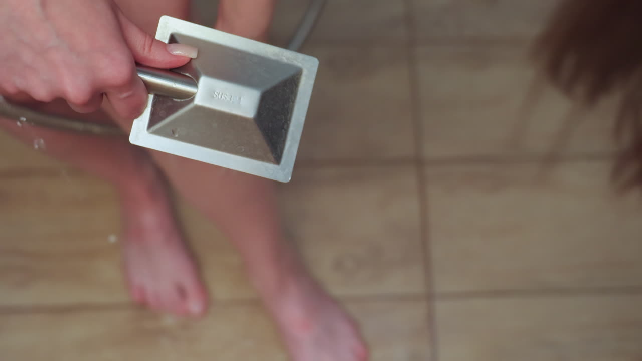 close up top down view of woman pouring water over leg with handheld shower in modern tiled shower room, towel wrapped around waist, water cascading over wet skin, capturing intimate self care ritual