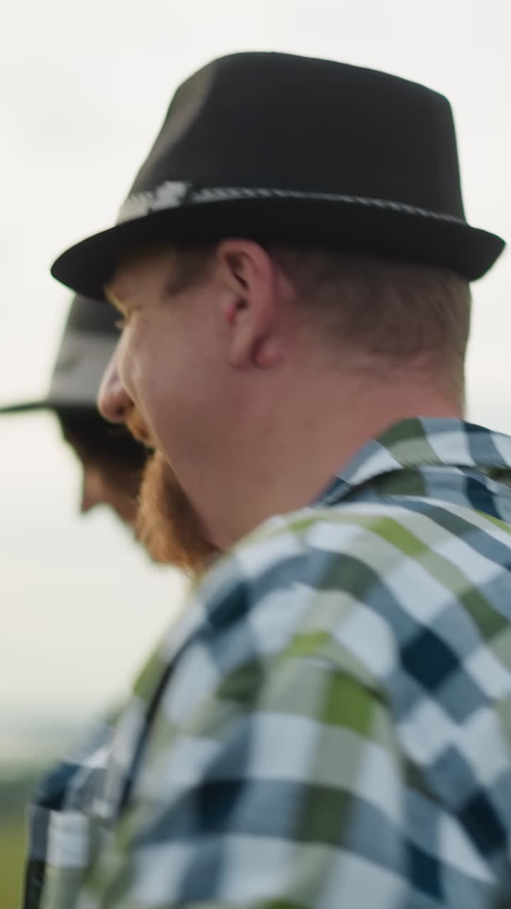 해가 지는 동안 잔디을 가로질러 달리는 즐거운 커플의 클로즈업, 웃음. 남자는 검은 모자와 체크 셔츠를 입고, 여자는 색 드레스를 입고 모자를 맞추고 있다.