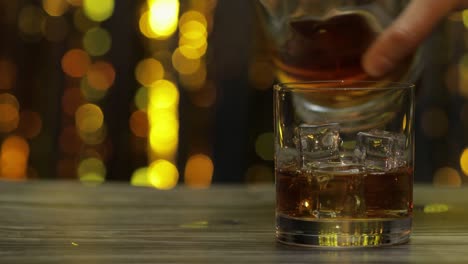 pouring of golden whiskey, cognac or brandy from bottle into glass with ice cubes. shiny background