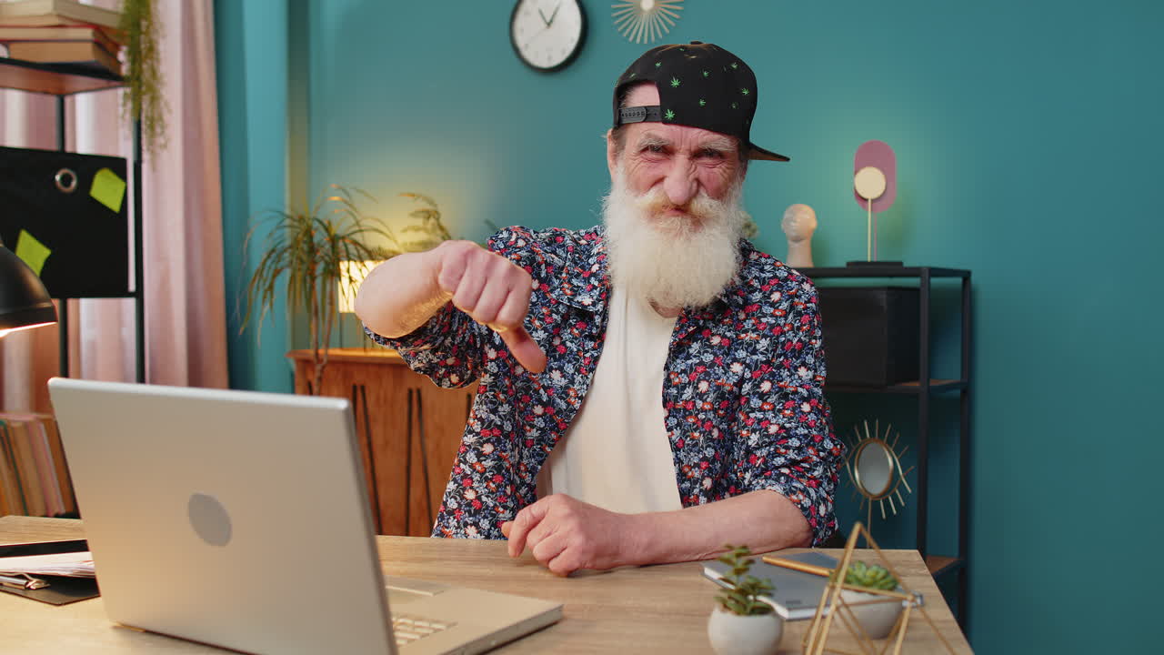 Elderly man working on laptop computer at home office showing thumbs down sign dislike disapproval