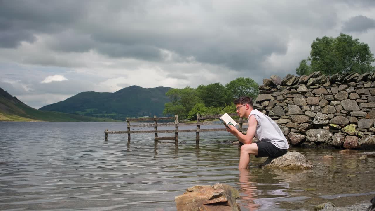 Man reads book with feet in the water sat on a rock in a lake. Lake District, UK