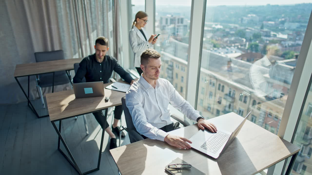 Young confident people working in the sunlit office. Male employees sitting at desks working on laptops and woman stands at window with phone. High angle view.
