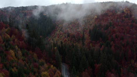 Winding-road-leading-into-misty,-moody-autumn-forest,-drone-crane-shot,-Romania