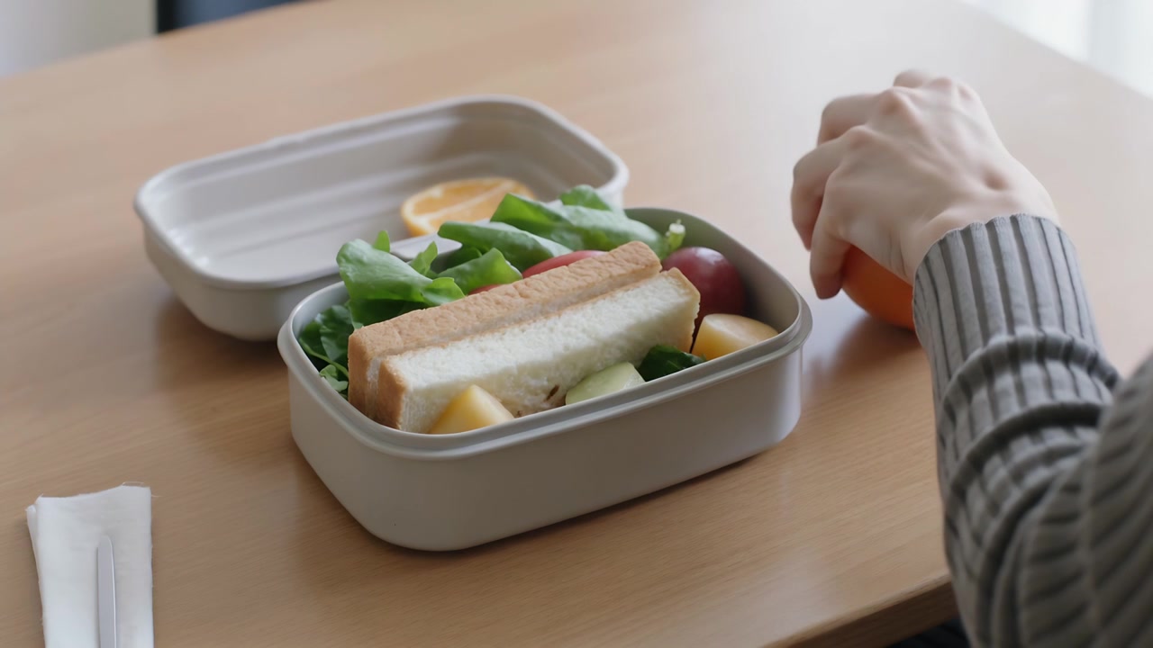 Von rechts kommende Hand in grauem Pullover legt Orange in Bento-Deckel beim Packen des Mittagessens am heimischen Tisch