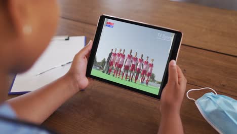 Composite-of-woman-sitting-at-table-with-face-mask,-watching-sports-event-on-tablet