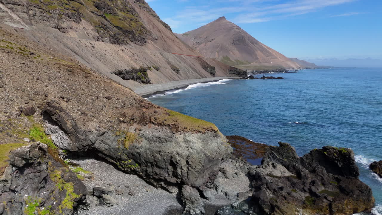 Aerial view of ocean waves hitting the rugged cliffside mountains of Iceland, revealing a winding coastal road, steep rocky slopes, and bright blue water stretching along the dramatic shoreline