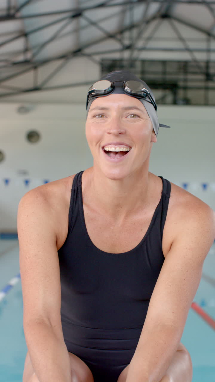 Vertical video: Smiling female swimmer in swimsuit and swim cap sitting by indoor pool
