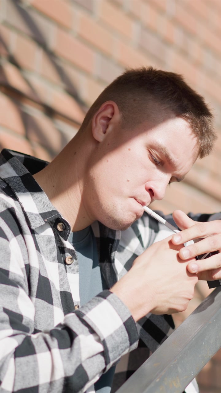 Close-up de jovem segurando e acendendo um cigarro enquanto veste uma camisa xadrez, com fundo de parede de tijolos. Luz natural suave realça as características do sujeito enquanto o homem realiza a ação, criando.