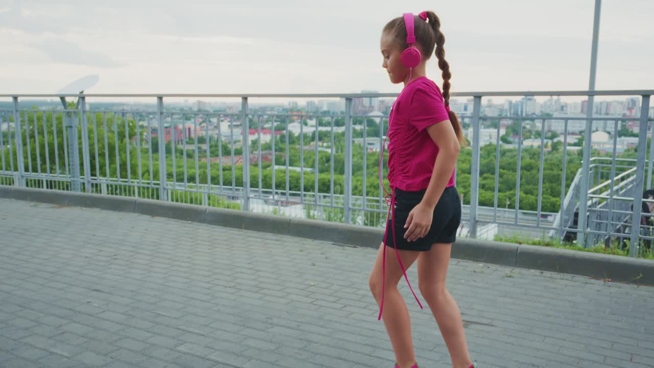 Young girl in pink headphones jumps rope joyfully in an urban environment, showcasing fun fitness and outdoor activity amidst a scenic backdrop of the city