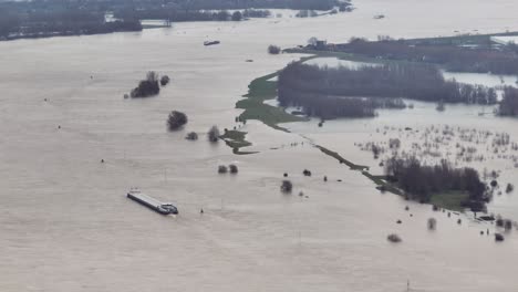 Große-Luftaufnahme-Eines-Schiffes,-Das-Auf-Dem-Fluss-Waal-In-Der-Nähe-Von-Gorinchem-In-Den-Niederlanden-Dampft,-Nachdem-Heftige-Regenfälle-Dazu-Geführt-Hatten,-Dass-Der-Fluss-über-Die-Ufer-Trat-Und-Bauernhöfe-Und-Gemeinden-überschwemmte