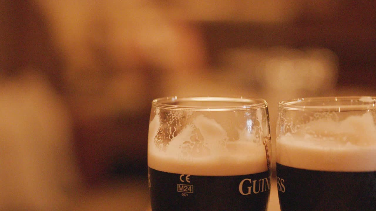 Two pints of dark stout clink together in a warmly lit pub, with a shallow depth of field and soft, blurred background