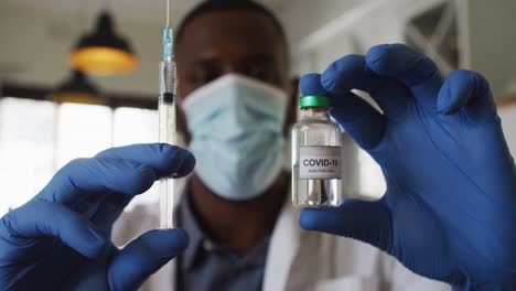 African-american-doctor-wearing-face-mask-holding-covid-19-vaccine-and-syringe-at-home