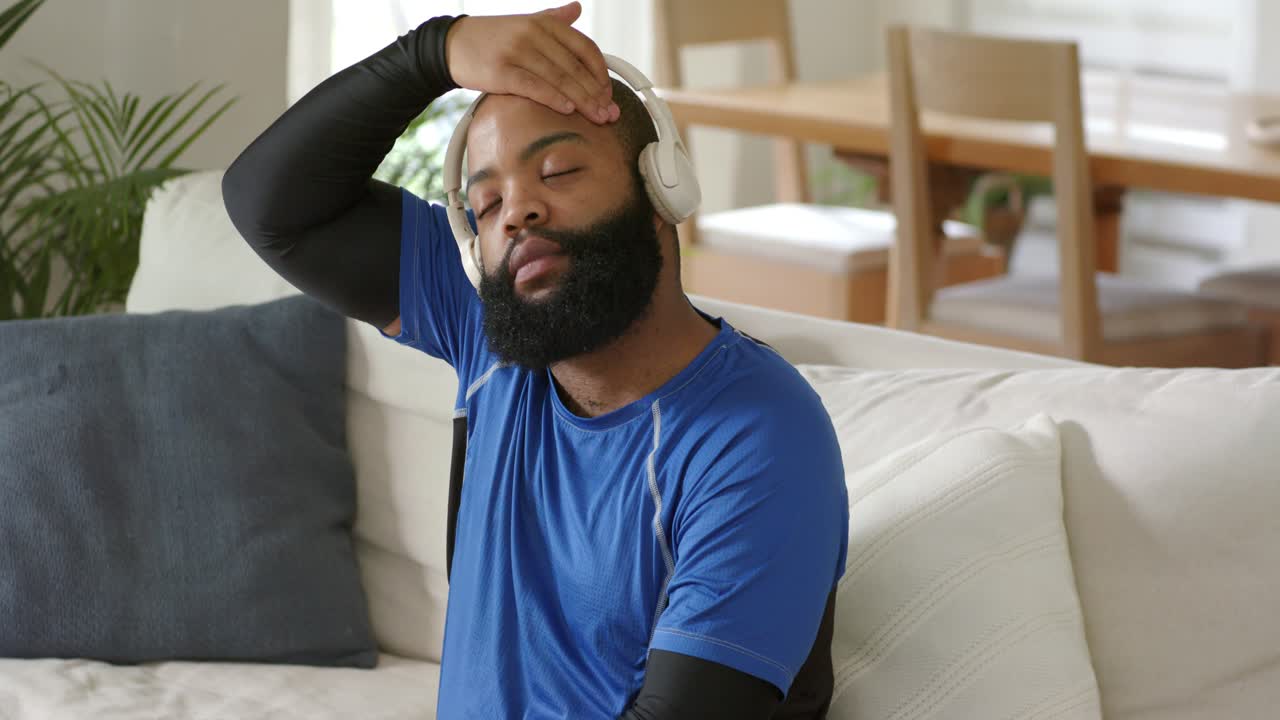 Adult African American man sitting on couch after closing eyes, adjusting white headphones for fit