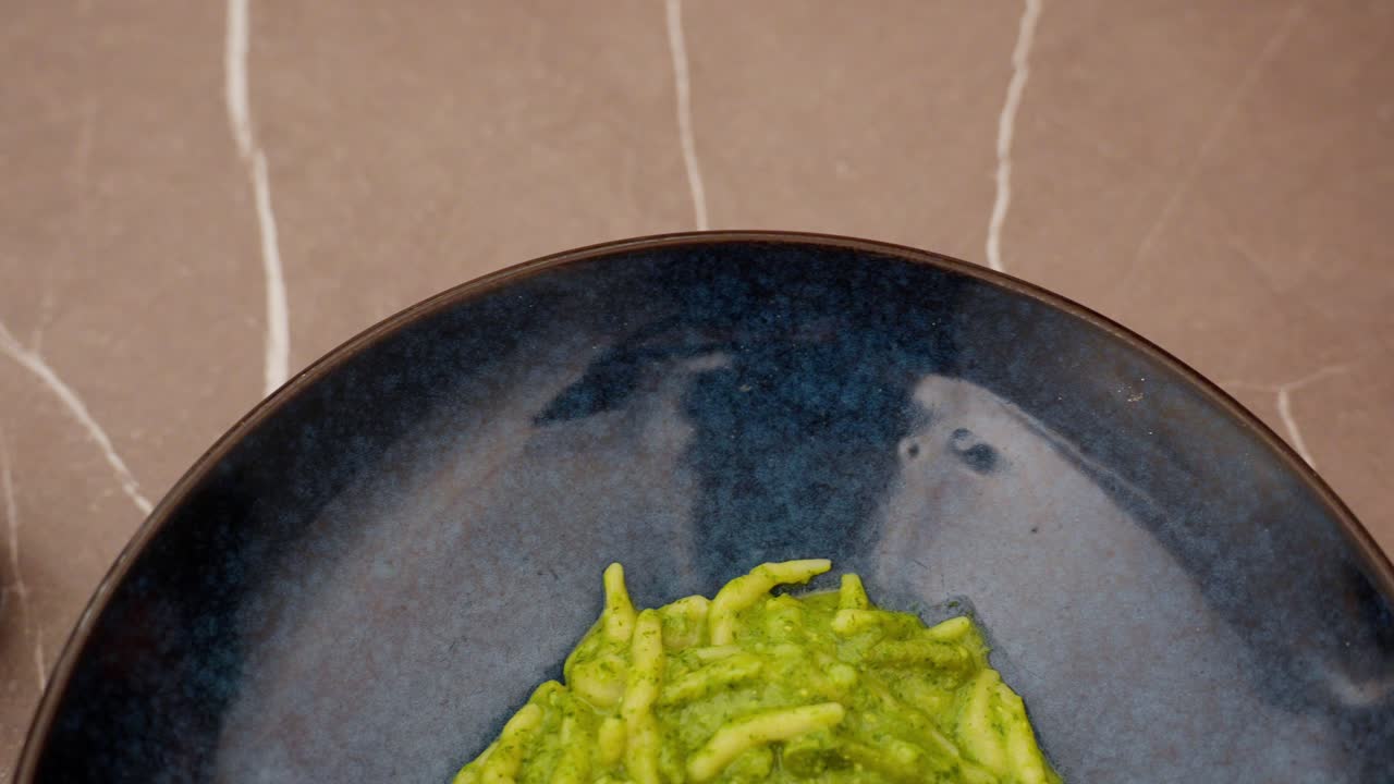 Pasta with pesto in a deep blue bowl, served with a glass of water on a light surface