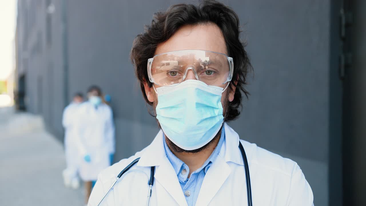 Portrait of Caucasian good-looking doctor wearing a medical mask, safety glasses and stethoscope looking at camera