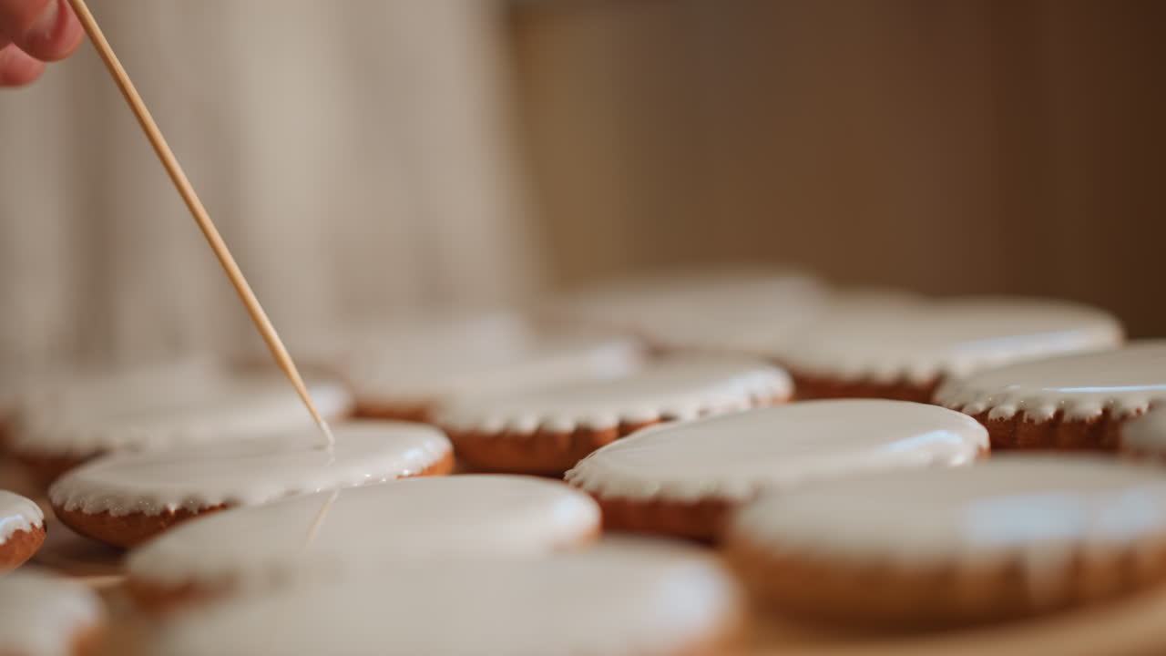 Iced cookies detailed with toothpick on wooden board, baker smoothing glossy icing, shallow focus, careful hands creating uniform patterns, soft warm window light, calm artisanal mood