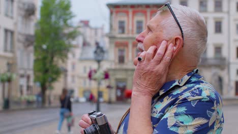 Senior-tourist-with-retro-photo-camera-listening-to-music-on-earphones-dancing-outdoors-having-fun