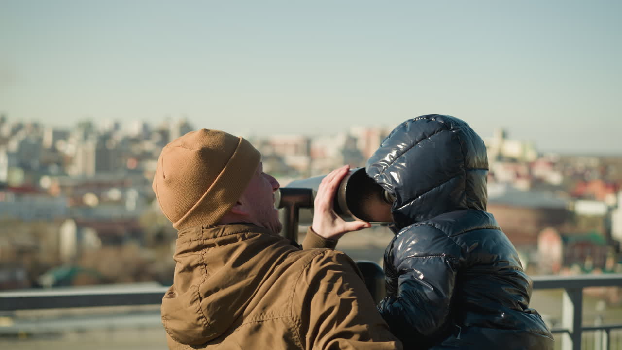 A father holds his son up to a telescope, guiding him looks through it, the father smiles, ensuring a steady view while the cityscape stretches out in the background