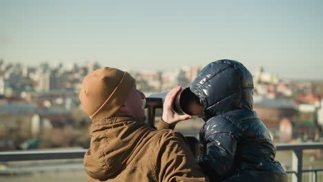 a father holds his son up to a telescope, guiding him looks through it, the father smiles, ensuring a steady view while the cityscape stretches out in the background