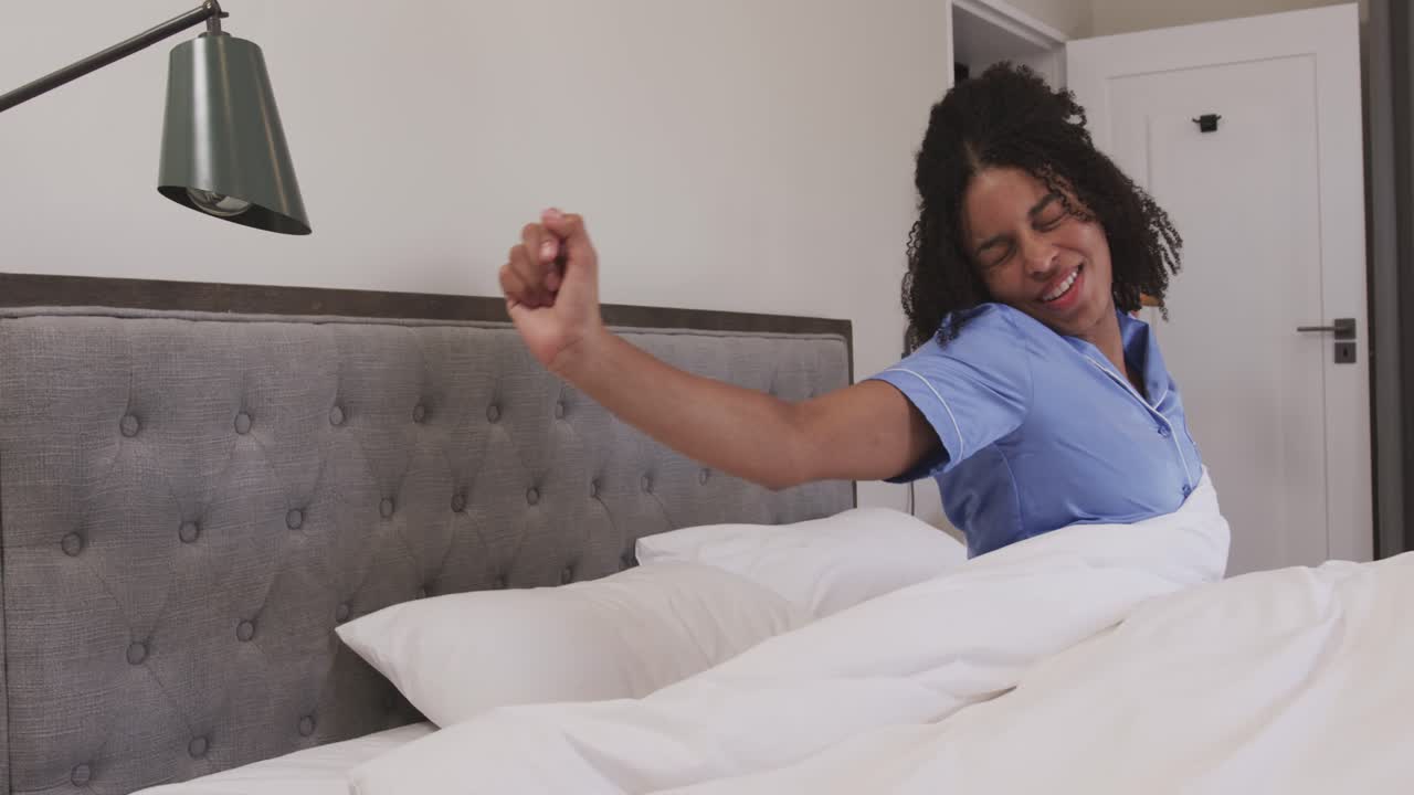 Donna afroamericana si sveglia, si siede a letto con un top del pigiama blu, tirando il piumone, e si stira.
