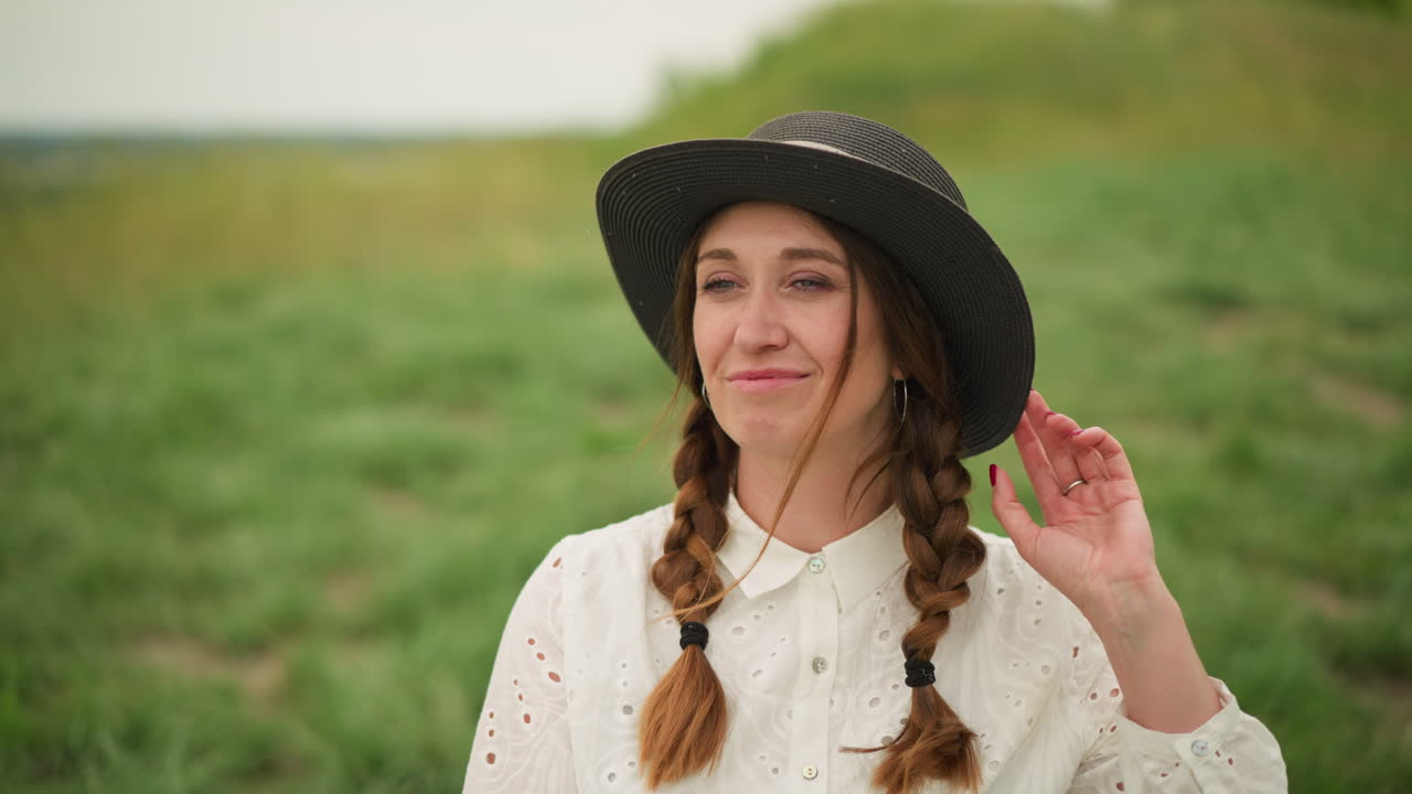 una mujer alegre con el cabello trenzado, con un vestido blanco y un sombrero negro, sonríe mientras está sentada casualmente en un campo verde exuberante
