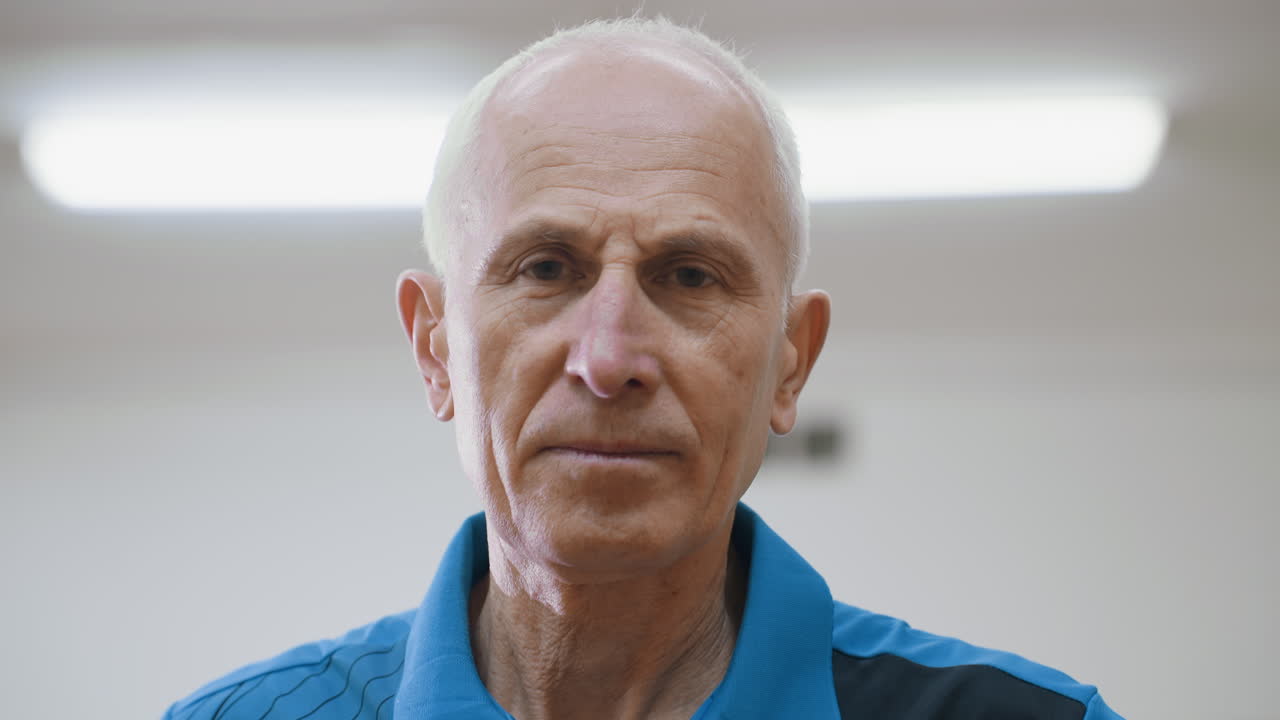 Primo piano di un uomo anziano con la mano sulla vita che alza la testa pensieroso in un ambiente interno, che mostra un'espressione calma, saggezza, maturità e uno stato d'animo riflessivo in un palazzetto dello sport