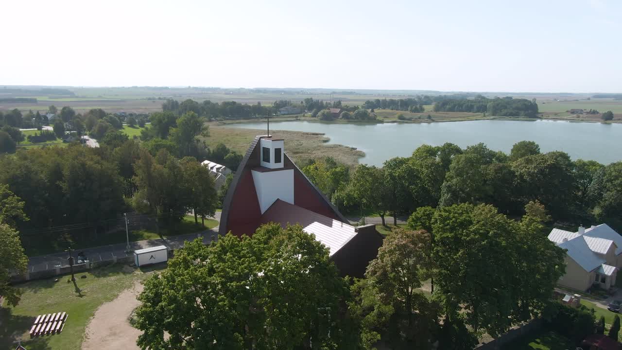 Drone passing over beautiful red roofed church and revealing beautiful small town and flatland with lake of Lithuania.
