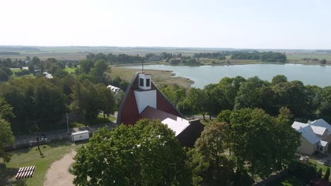 drone passing over beautiful red roofed church and revealing beautiful small town and flatland with lake of lithuania.