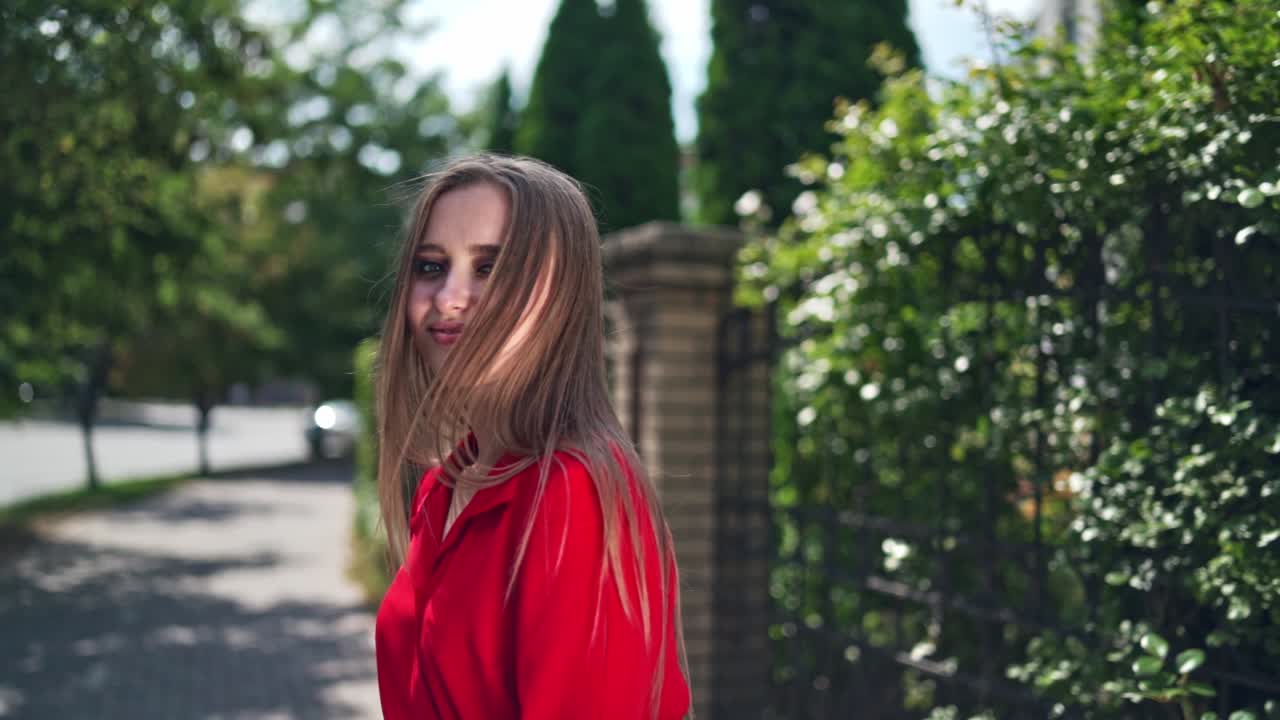 Passionate young girl posing to camera. Beautiful young model in red dress with long hair on the green trees background. Sexy woman turning around herself joyfully.