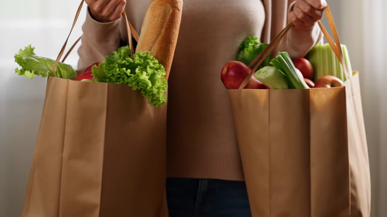 Mujer sosteniendo bolsas de papel para compras, llenas de lechuga, manzana, pepino y baguette. Torso de compradora casual enmarcado