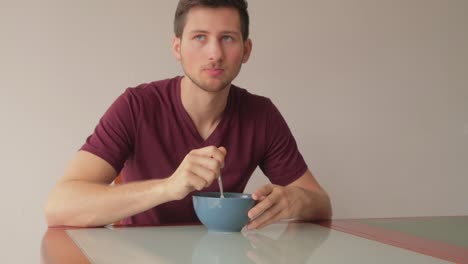 young man sitting at table eating cereals with neutral expression, still shot indoors in minimalist setup