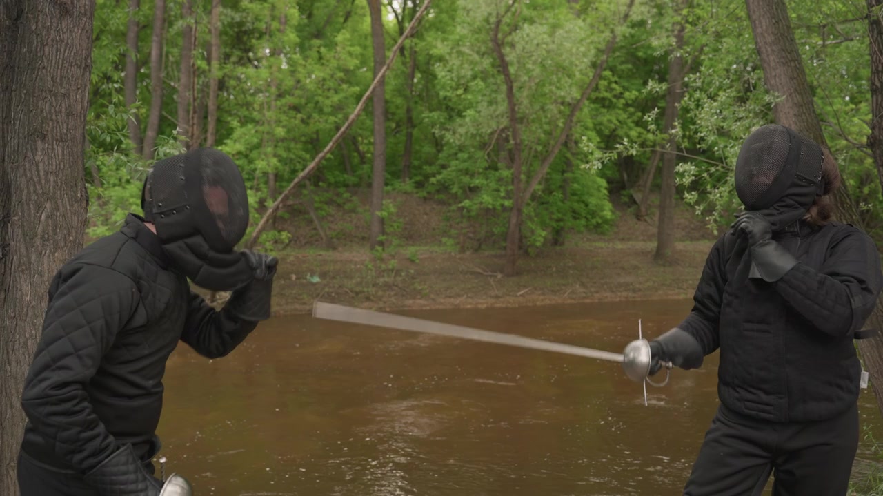 Two people in black fencing gear practice swordplay by river adjusting their masks and engaging with each other. scene features natural greenery surrounding water body creating outdoor