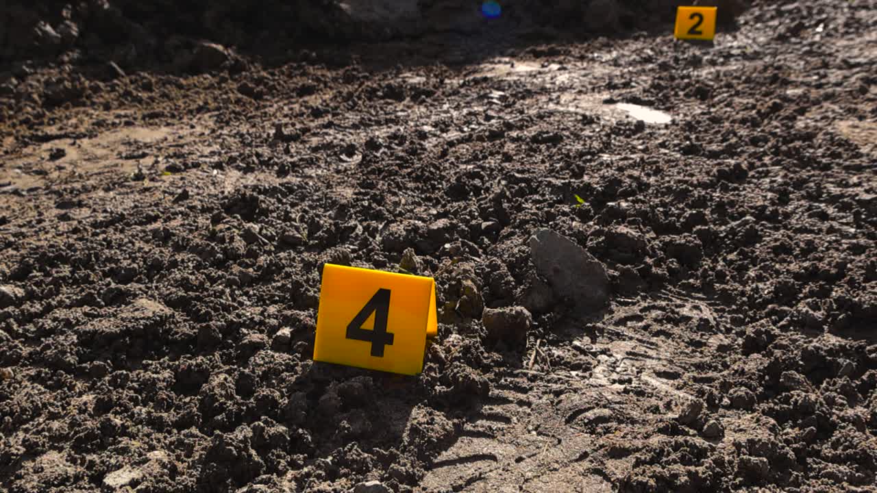 Close up view orbiting and spinning around a yellow colored police crime scene marker placed next to a boot or shoe footprint in a damp and wet muddy road at rural countryside during a sunny day