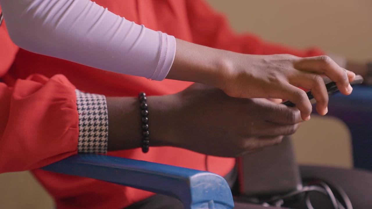 Hands of black mother and daughter, closeup on fingers with bracelet and red sleeve over blue armrest, device exchange and gentle touch, tactile connection and trust, domestic detail with screen focus